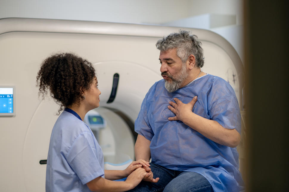 An older man with a beard, wearing a medical gown, hand on his chest, speaking to a woman radiology technician in scrubs