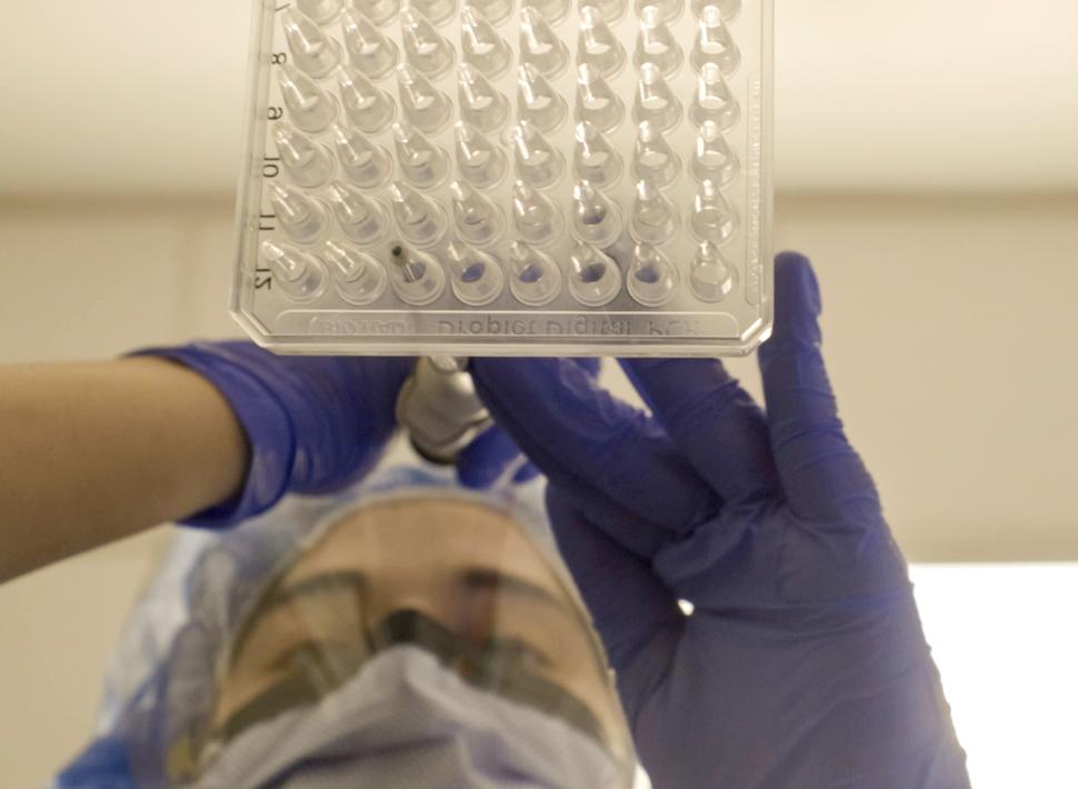 A picture from below of a lab tech wearing a mask, safety glasses, and blue gloves dropping materials from a pipette into a 64-well plate.