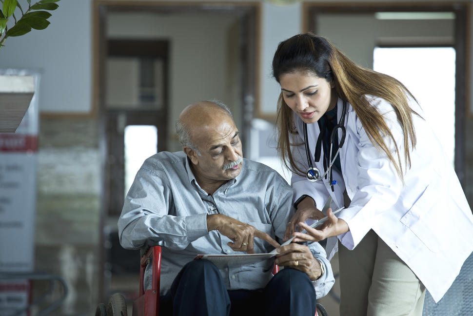A patient points at and looks toward a folder in his lap while a doctor leans in next to him, gesturing and looking toward the folder as well.