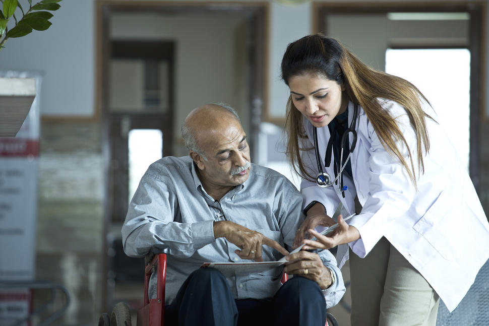 Un paciente mira y señala la información en una carpeta que tiene en su regazo, mientras una profesional de la salud se reclina hacia él y observa la misma información.
