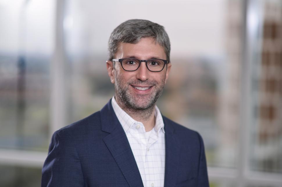 Man with grey and dark hair wearing glasses, a blue suit jacket, and a white dress shirt.