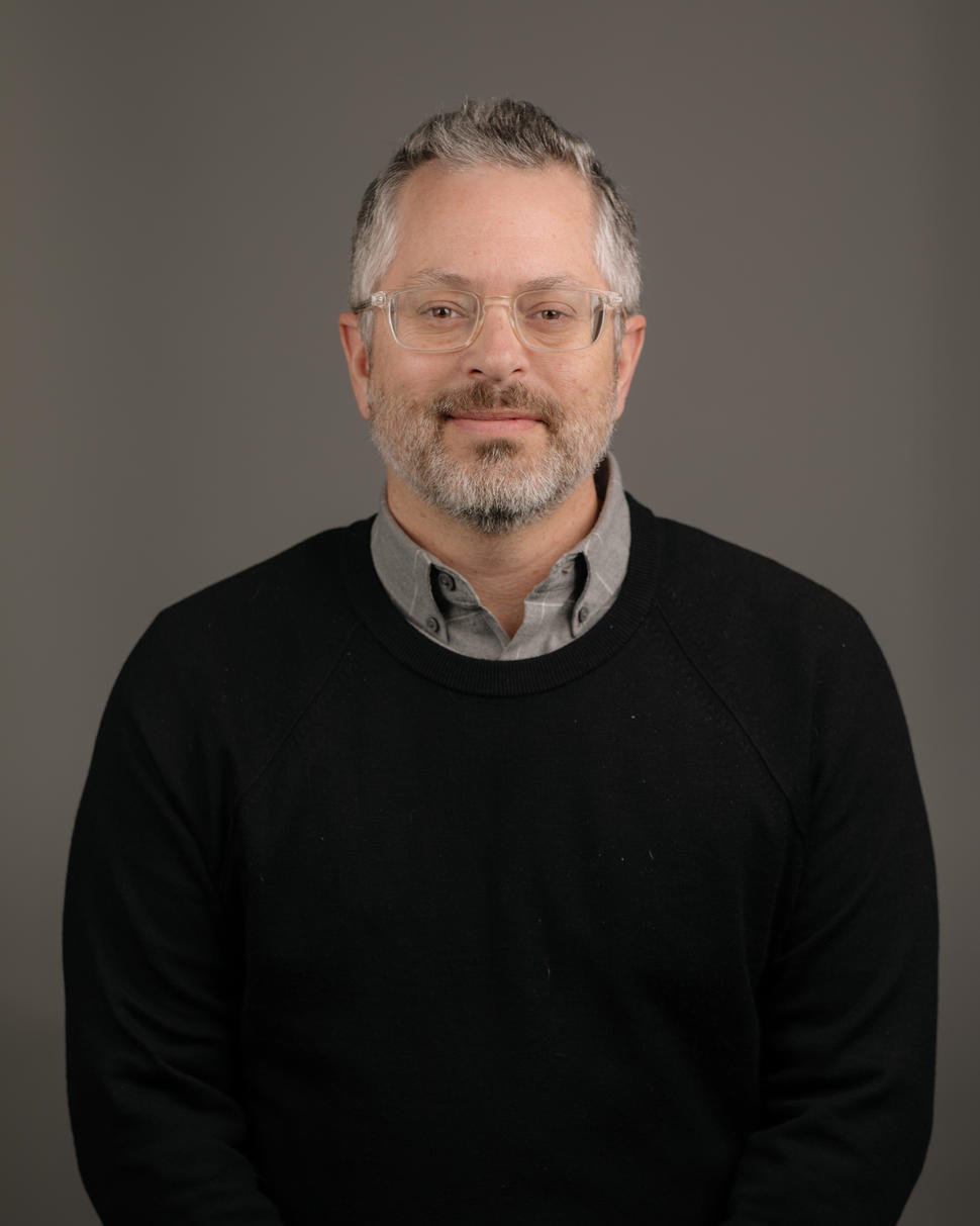 Bearded man wearing clear glasses and a black sweater over a grey collared shirt