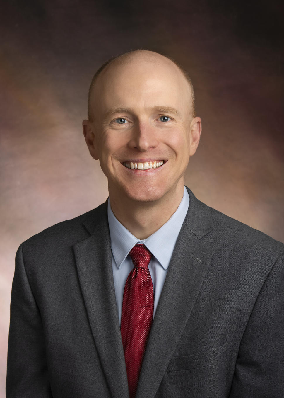 Man in grey suit, light blue shirt, and red, dotted tie smiles at the camera