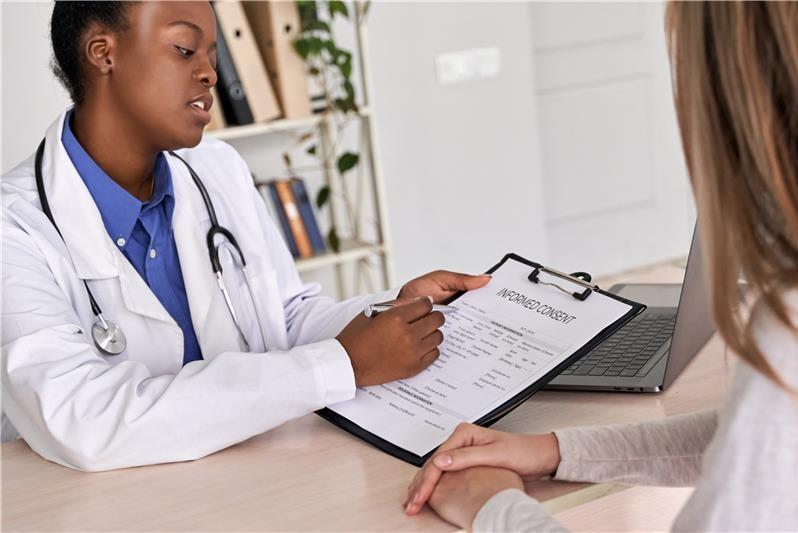 Clinician and patient sitting across from each other at a desk. The clinician is holding a clipboard with a paper that reads Informed Content at the top. The patient has her hands folded together as she listens to the clinician explain the document and signature requirements.