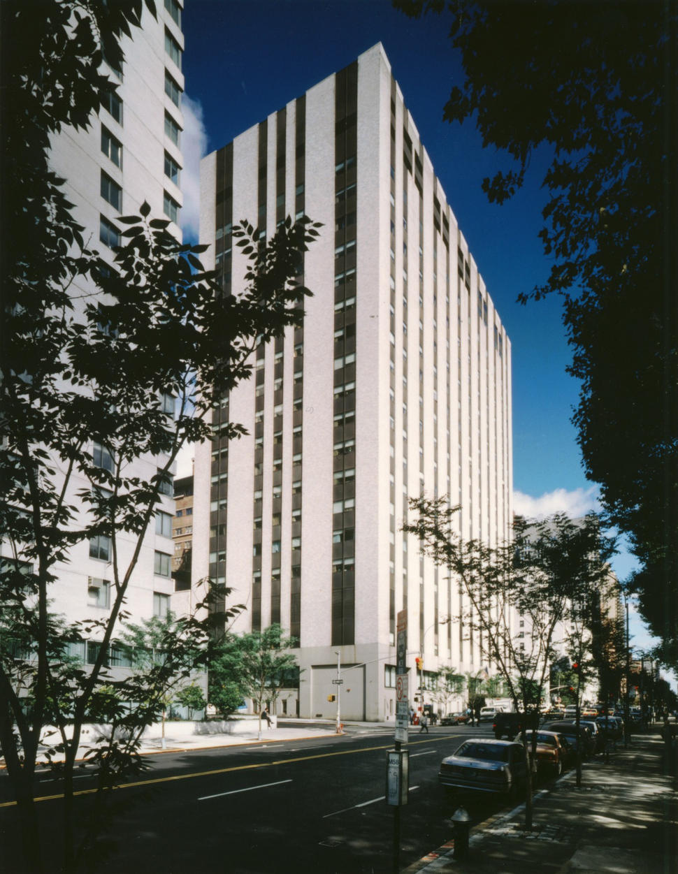 External view of Memorial Sloan Kettering Cancer Center. The building has a modern design, featuring vertical white and dark panels. The structure is set against a clear blue sky, framed by trees and a street lined with parked cars.