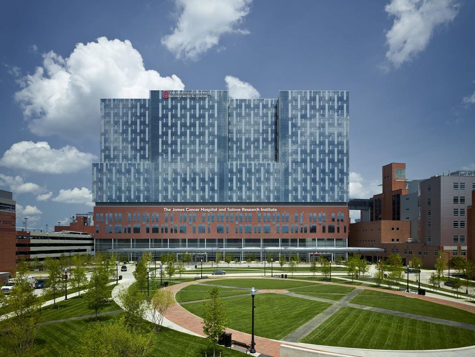 An exterior photo of the Ohio State University Comprehensive Cancer Center. The building is modern and mostly glass for the top 2/3s; the bottom is brown with windows. Smaller buildings can be seen on the left and the right; in front of the cancer center, a large green park with trees can be seen. The sky is blue with large fluffy clouds.