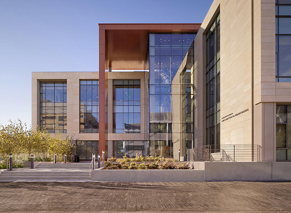 An exterior photo of the Stanford Cancer Institute. The building has hard edges and large glass panel windows that take up a majority of the exterior. The parts of the building that aren’t glass are greys and warm oranges. There are steps seen on both the left and right of the building, along with trees and bushes throughout. The sky is blue and cloudless. 
