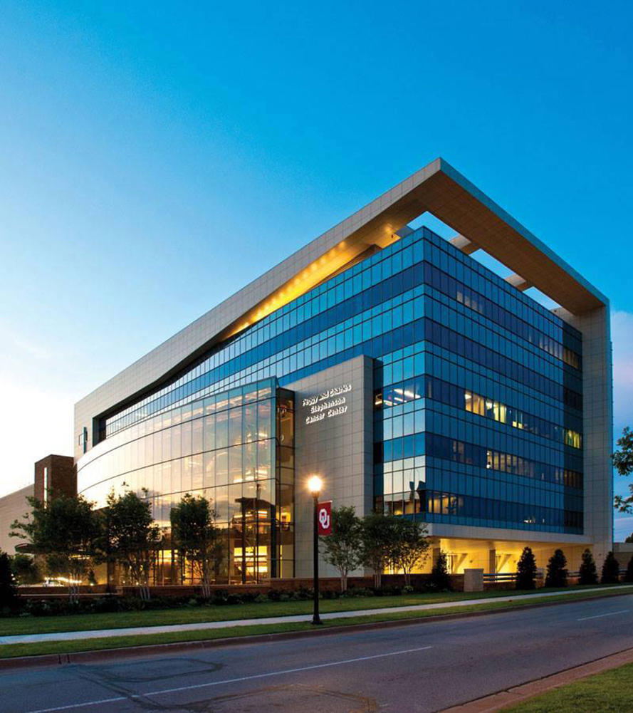 An exterior photo of the Stephenson Cancer Center. It is a modern building that is mostly made of glass windows. A large angular piece of the building at the top frames the lower floors. A row of trees runs along the length of the building. The sky is blue and the light hits the building’s windows in a way to suggest sunrise or sunset.