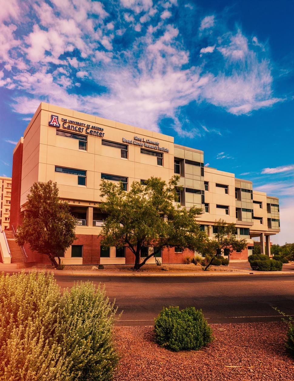 Exterior of the University of Arizona Cancer Center, a multi-story beige building with rows of windows, desert landscaping, and a partly cloudy blue sky.