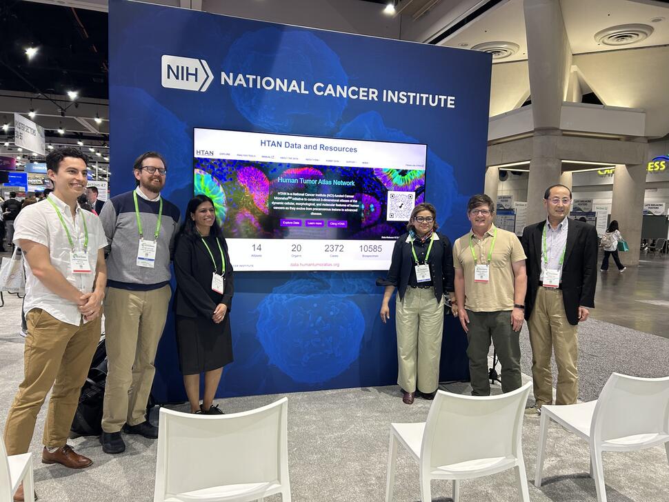 Photo of 6 people involved in HTAN standing next to a slide that says the Human Tumor Atlas Network (3 people per side)