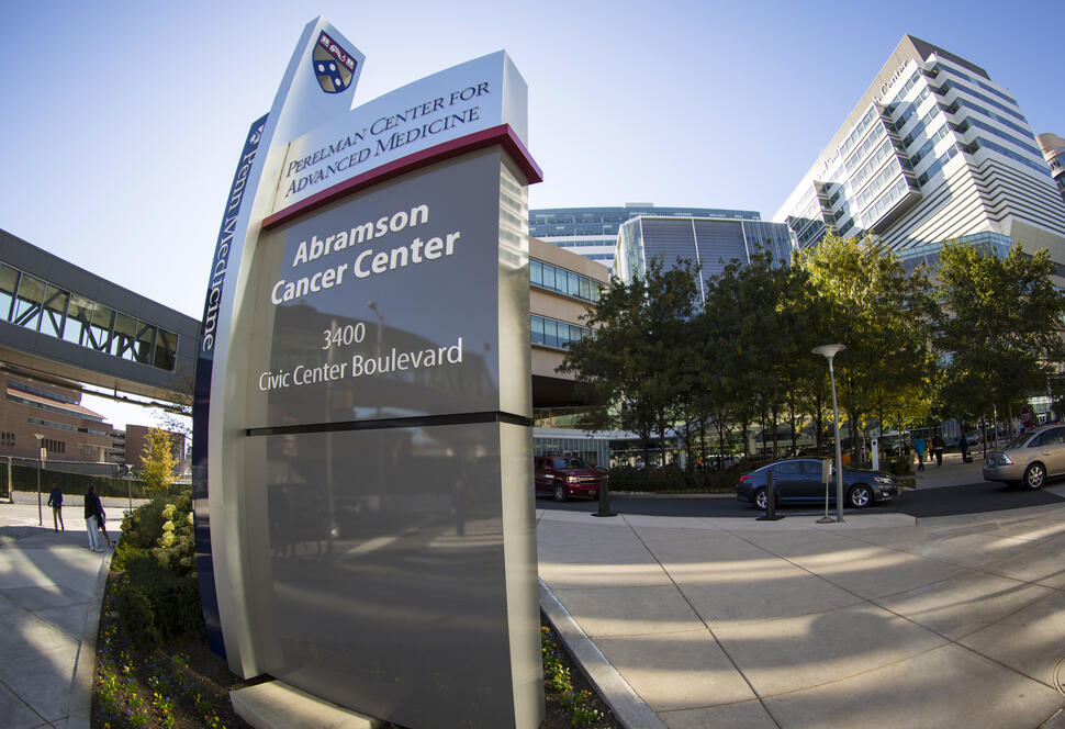 Abramson Cancer Center sign at the University of Pennsylvania with surrounding buildings, cars, and pedestrians on a city street.