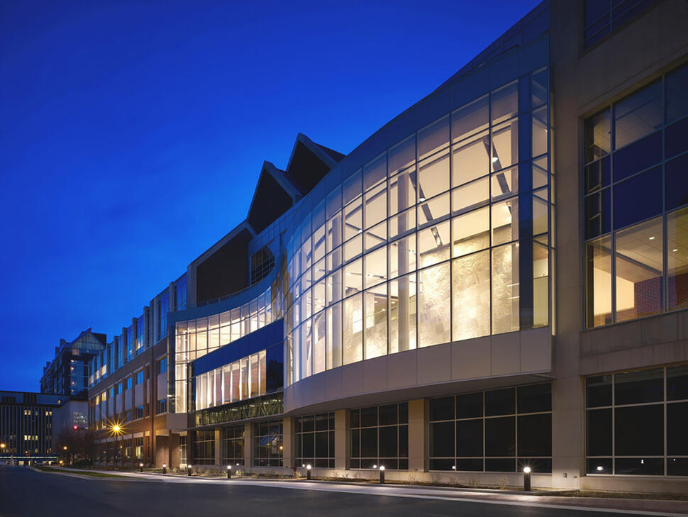 Indiana University Melvin and Bren Simon Cancer Center with a curved glass facade illuminated at dusk along a quiet street.