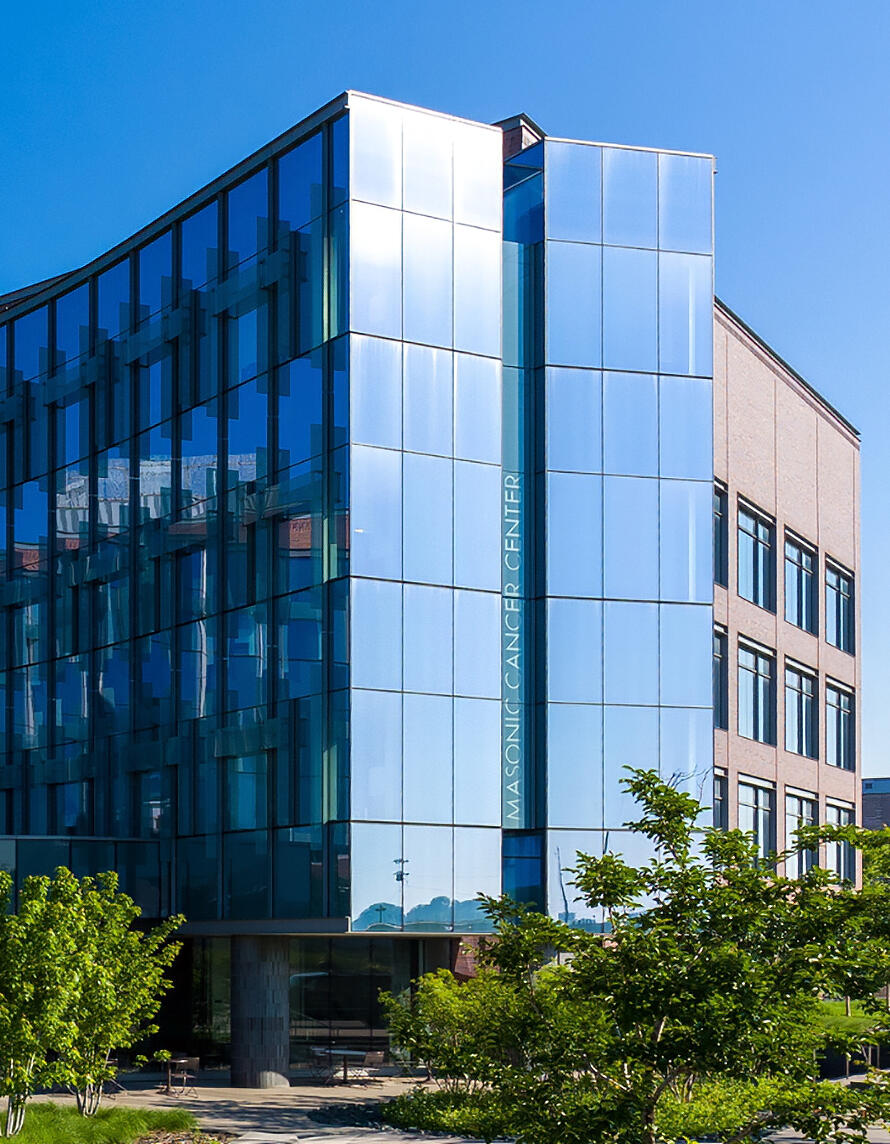 Masonic Cancer Center at the University of Minnesota, a modern glass and metal building with reflective panels and trees in front.