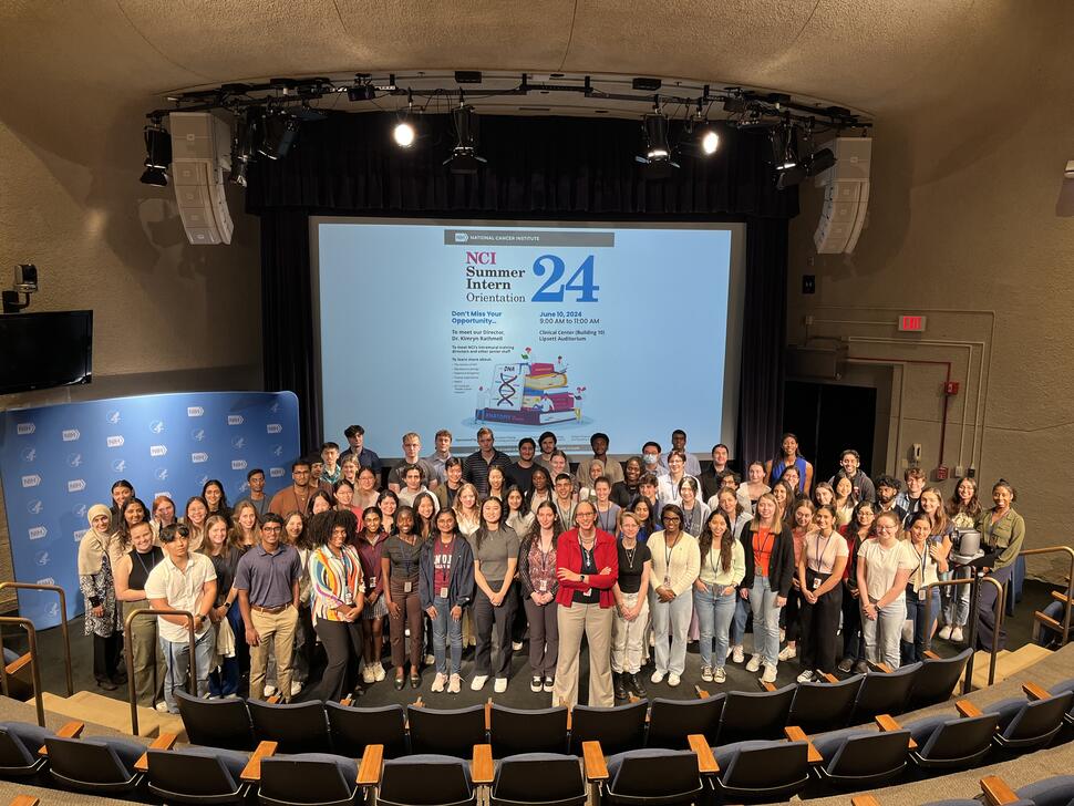 Group photo of 2024 National Cancer Institute (NCI) summer interns gathered in an auditorium with then-NCI Director Dr. Kimryn Rathmell, standing at center front in a red blazer. The group stands onstage beneath a large screen displaying “NCI Summer Intern Orientation 2024” at the NIH Clinical Center in Bethesda, Maryland.