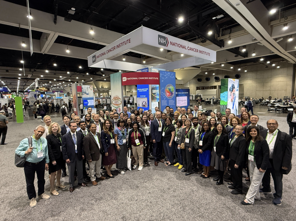 About 70 professionally dressed people smiling at the camera and standing in a semicircle in front of the NCI exhibit booth.