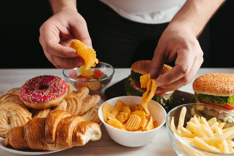 Hands reaching for ridged potato chips from a bowl surrounded by burgers, fries, pastries, candy, and a frosted doughnut.