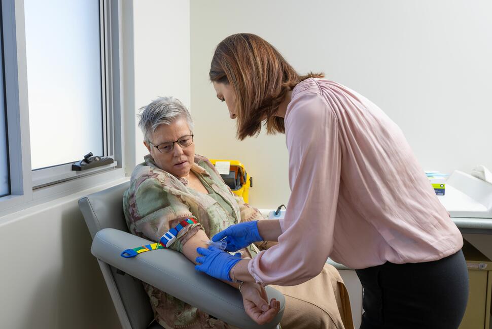 Health care provider wearing blue gloves performs a blood draw on a seated patient in a clinic exam room.