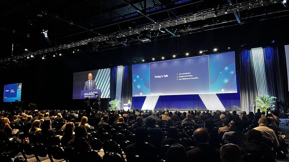 Dr. Anthony Letai stands at a lectern on a stage and addresses attendees filling rows of seats in a large conference hall.