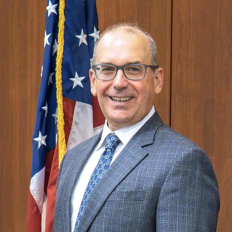 NCI Director Dr. Letai standing in front of a flag and brown wood background.
