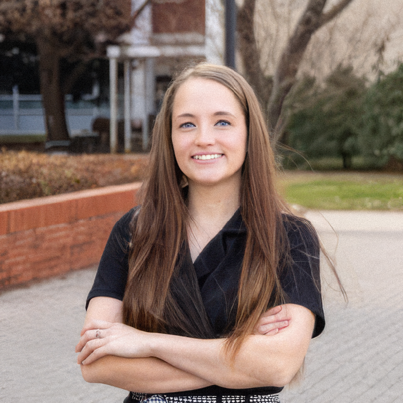Gabrielle smiles at the camera with her arms crossed, wearing a black top and standing outdoors near a brick building and landscaped lawn.