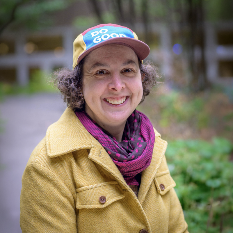 Olivia smiles at the camera, wearing a yellow jacket, patterned scarf, and multicolored cap with the words “DO GOOD.”