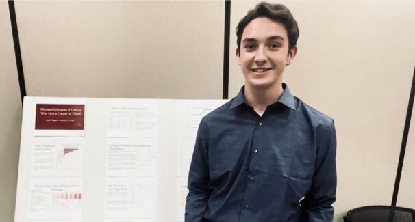 Jacob Hogan wearing a blue button down shirt in front of a poster on Human Lifespan if Cancer Was Not a Cause of Death."