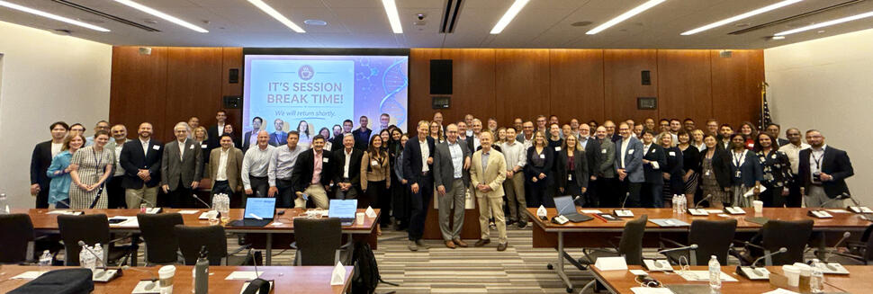 A large group of workshop attendees poses for a photo in a large meeting room. A screen behind them reads, “It’s session break time! We will return shortly.”