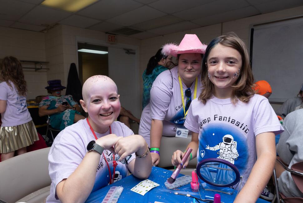 Three young people wearing Camp Fantastic shirts gather around a table covered with makeup, glitter, and craft supplies. Two smile toward the camera while another, wearing a bright pink cowboy hat, stands behind them.