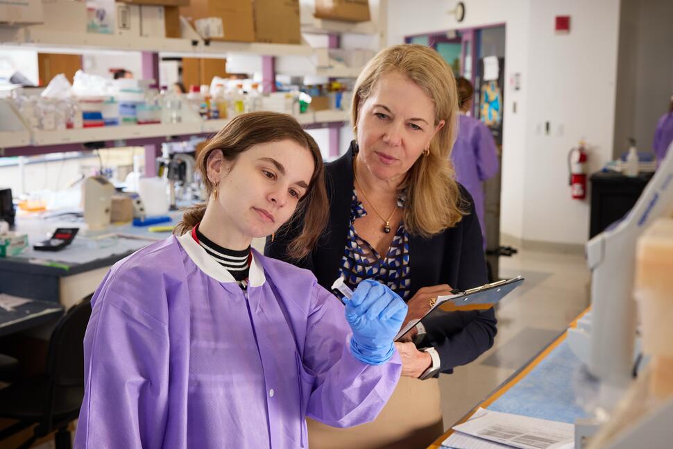 A laboratory researcher examines a sample while a colleague observes and takes notes.