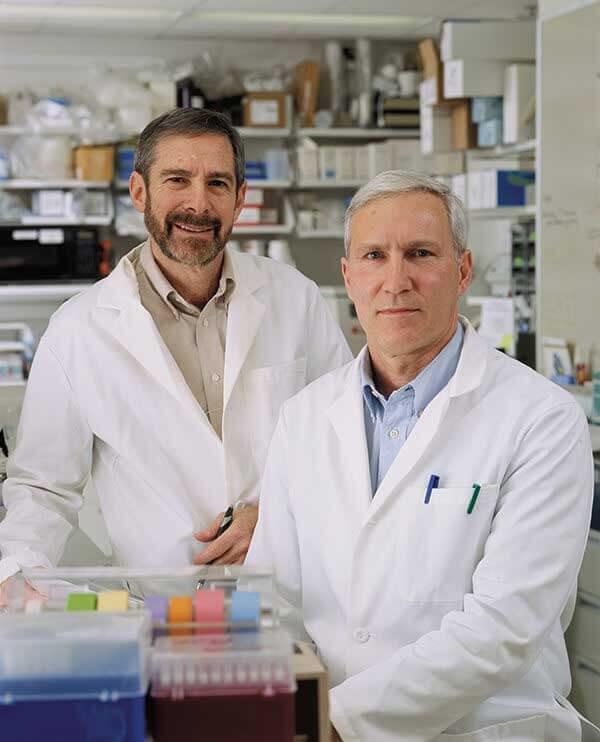Dr. Douglas Lowy and Dr. John Schiller, standing side by side in white lab coats in a research laboratory setting with equipment and shelving visible in the background.