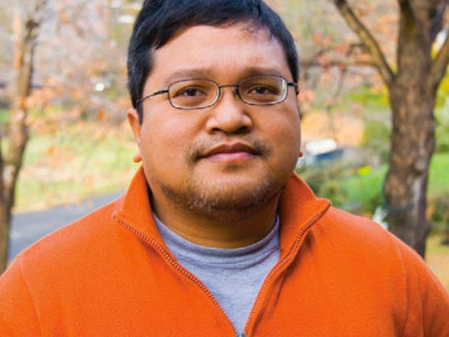 An Asian man, who has dark hair and glasses, looks at the camera. He’s wearing an orange quarter zip sweater over a grey shirt.. In the background, there are tree trunks and greenery, indicative that he may be in a park. 