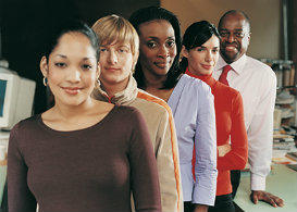 A group of five people, all adults, stand in a line. There are three women and two men. They are all dressed in business appropriate clothing.The background features computers and desks, indicative that they are in an office.