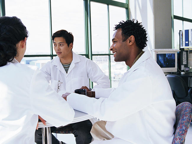 Three people in lab coats - two men and one woman - sit at a small table near a large window. One of the men is smiling at his colleague. In the corner is a desk with a computer and binders.