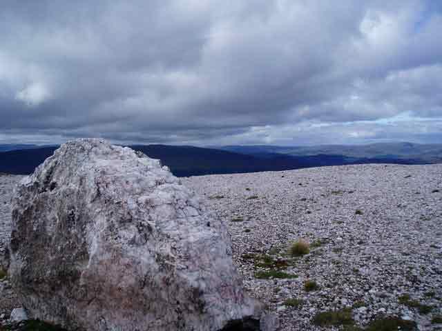 A large chunk of quartz sits on top of rocky terrain. In the background, there’s a cloudy sky and dark mountains.