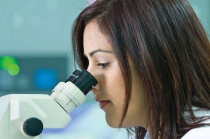 A close-up photo of a brown-haired woman looking into a microscope. The background is blurry and you can make out some glowing lights.