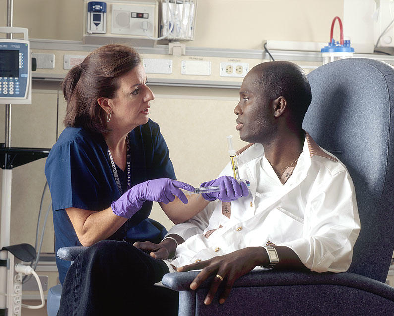 A nurse administers chemotherapy to a patient. Both are seated. The nurse has shoulder-length brown hair and wears dark teal scrubs and purple gloves. She is holding a syringe that is held against the patients chest. The patient is wearing casual clothing and he looks at the nurse.