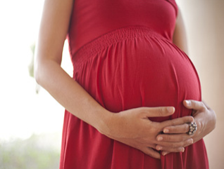 A close-up of a woman’s arms holding her pregnant belly. She is wearing a red dress.