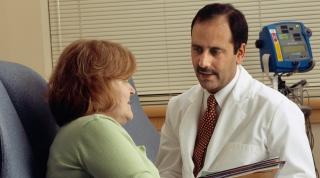 Doctor in a white coat and tie having a consultation with a female patient in a green shirt in a clinical examination room.