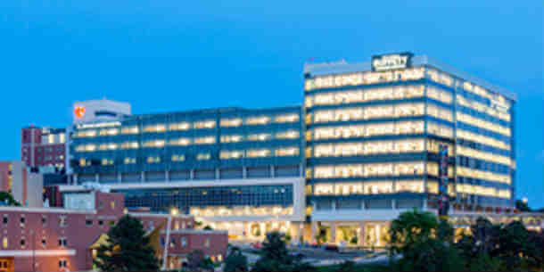 An exterior photo of the Fred and Pamela Buffett Cancer Center. The building is modern and has a larger horizontal section on the left connected to a squarish section on the right. Larger windows are seen in the right section than in the left. In the foreground, tops of smaller buildings and treetops can be seen. The sky is a dark blue and the windows are lit with bright yellow, indicating the photo was taken after the sun has set.