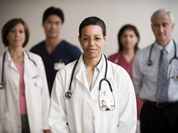 Five medical professionals - two men and three women - look at the camera with neutral expressions. A female doctor, wearing a lab coat, glasses, and stethoscope, is in focus in the foreground; the other four people are out of focus in the background. Two of the people - one man and one woman - in the background wear nurses scrubs. The other man and woman have stethoscopes and business professional clothing, indicating they are doctors.