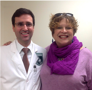 Dr. Christian Hinrichs and Susan Scott stand looking at the camera and smile. Dr. Hinrichs has short dark hair, is wearing glasses, a white coat, and shirt and tie. Susan has short, dirty blonde hair, is wearing sunglasses on top of her head and a large purple scarf and top.