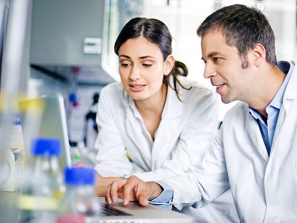 Two scientists wearing lab coats are collaborating at a computer in a laboratory setting