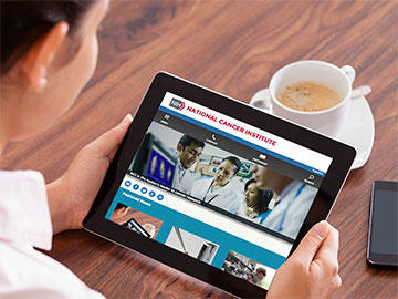 A person, seen from behind and wearing a white shirt, holds a tablet horizontally which displays the National Cancer Institute website. Behind the tablet is a cup of coffee and a cell phone is also nearby. 