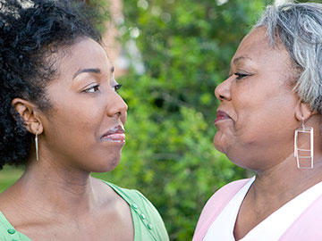 A mother and daughter look at each other with happy, bemused expressions. Both women are black. The mother’s hair is grey, whereas the daughter’s hair is black. Both wear casual clothing. In the background are bushes, indicating an outdoor environment.