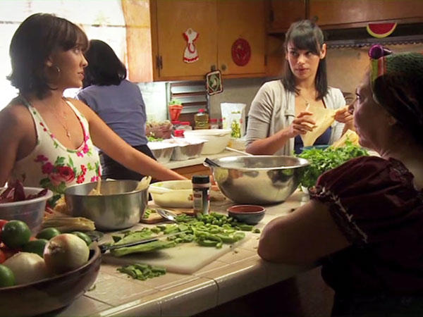 Two younger women in casual clothing look at each other while they prepare food. In the foreground, and also in shadow, an older woman looks on. Another woman is in the background with her back to the camera. The kitchen island in front of the two younger women and older woman has food and mixing bowls on it.