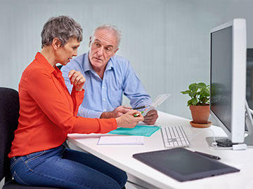 Woman and man seated in front of desktop computer and viewing paperwork.
