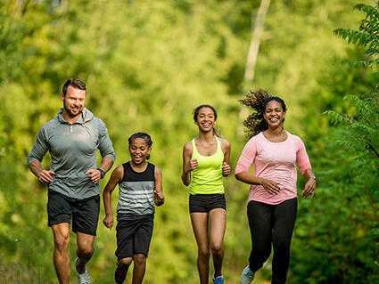 Father, mother, and two daughters run together surrounded by park greenery.