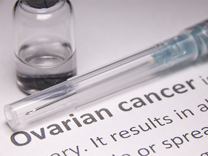 A close-up of a syringe and medical bottle containing clear liquid. These two items are sitting on a piece of paper that shows the definition of ovarian cancer. The words “ovarian cancer” are bold; the rest of the text is mostly cropped off the image, though what is shown indicates it is a definition of ovarian cancer.