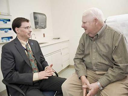 A doctor and a patient, both sitting, speak to each other. The doctor is middle aged with short dark hair, glasses, a grey suit and tan shirt with patterned tie. He’s sitting in a chair. The patient is an elderly man in a green button up shirt and khakis with white hair; he sits on an exam chair. A sink, cabinets, and gloves are in the background.
