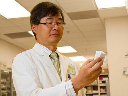 A pharmacist with short black hair and glasses stands in a pharmacy examining a boxed drug.